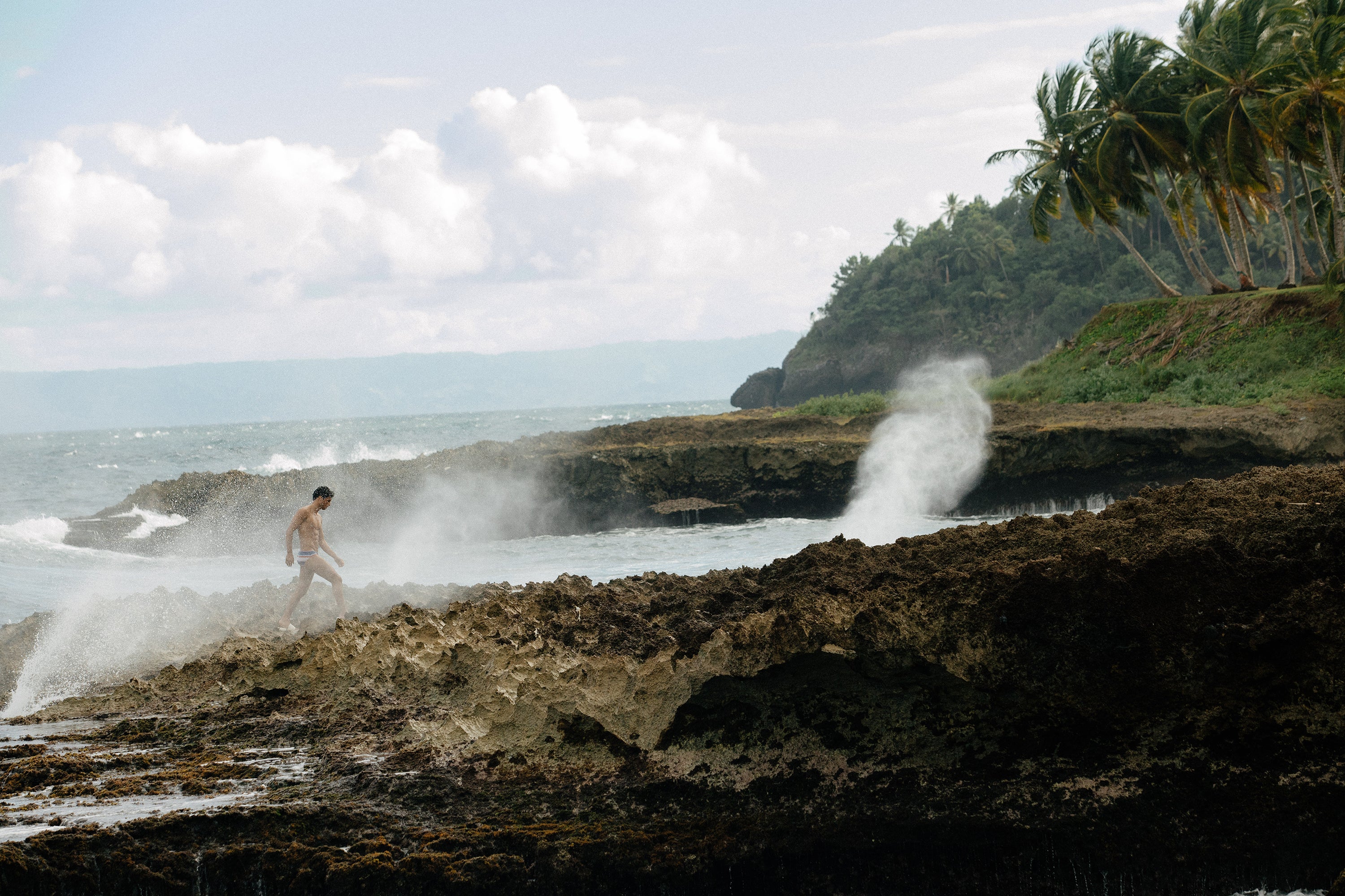 Landscape imag. Model walks on the beach next to scenic tidal pool spray. He Wear Parke & Ronen Meridian Swim biref.
