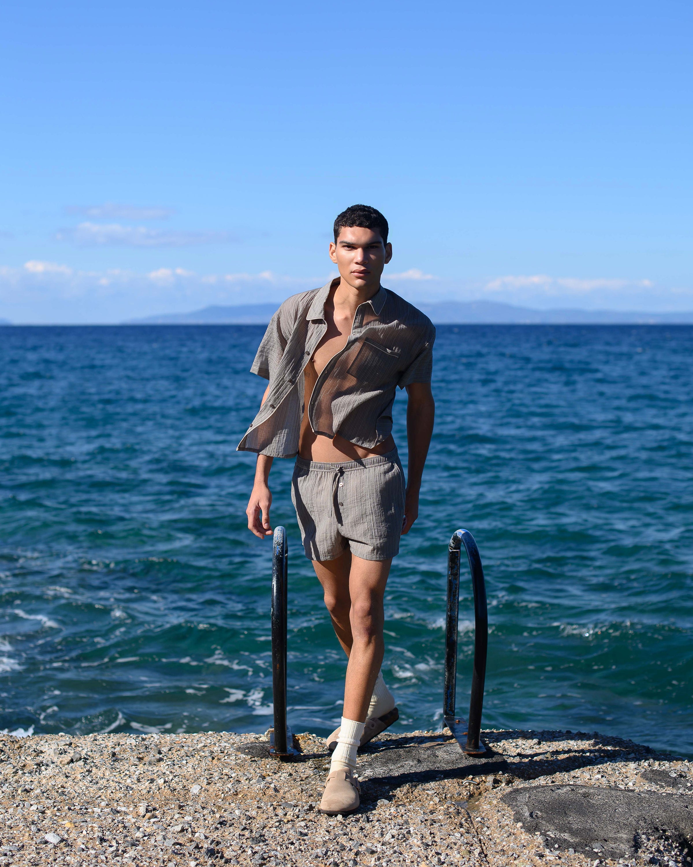 Male model walks on pier wearing Parke & Ronen khaki cropped shirt and matching drawstring shorts