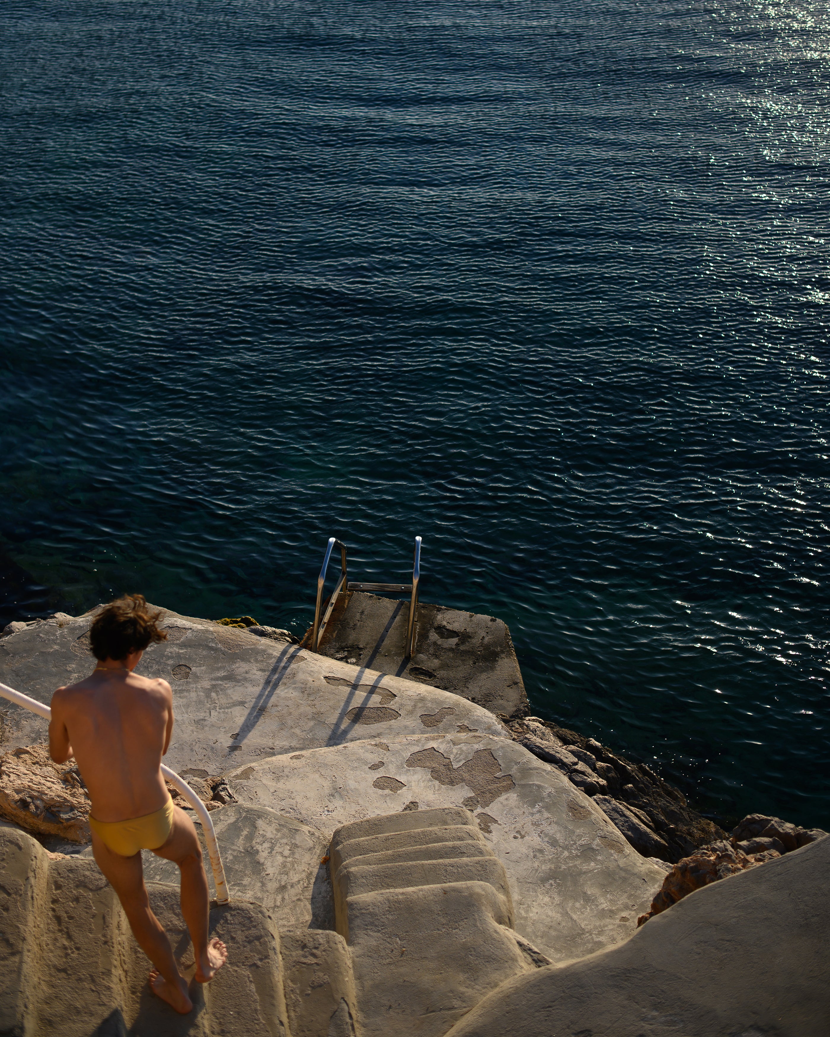 Parke & Ronen Spring 2024 Campaign Image. Male model stands seaside in gold Blade swim brief.